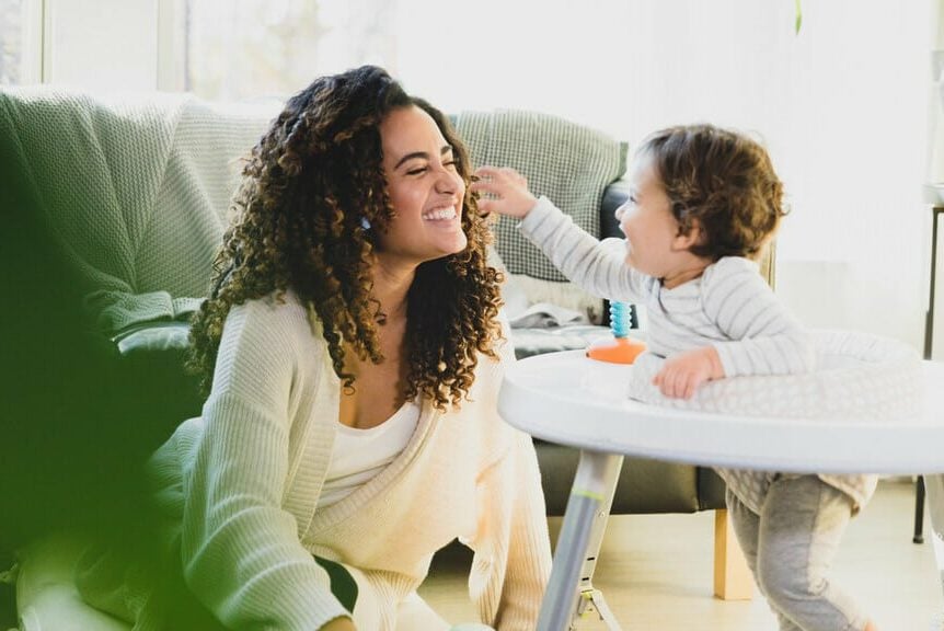 An au pair plays with some toys with her host child, an infant, in the living room. Au pair requirements for the United States.