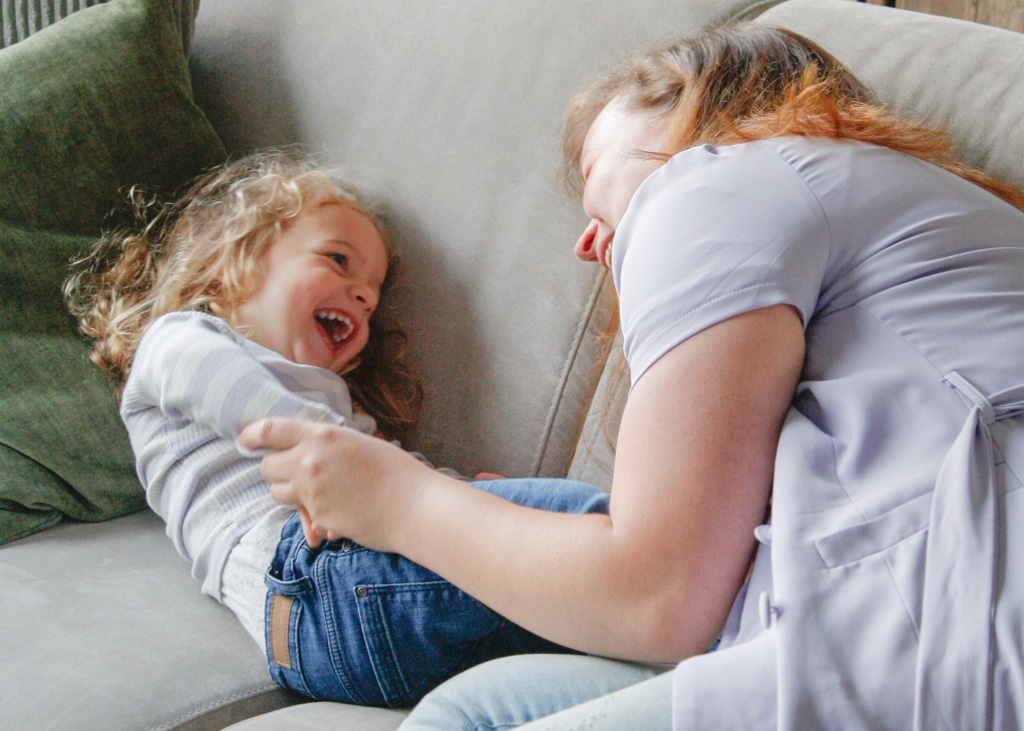 An au pair plays with her laughing host child on the couch in their home.
