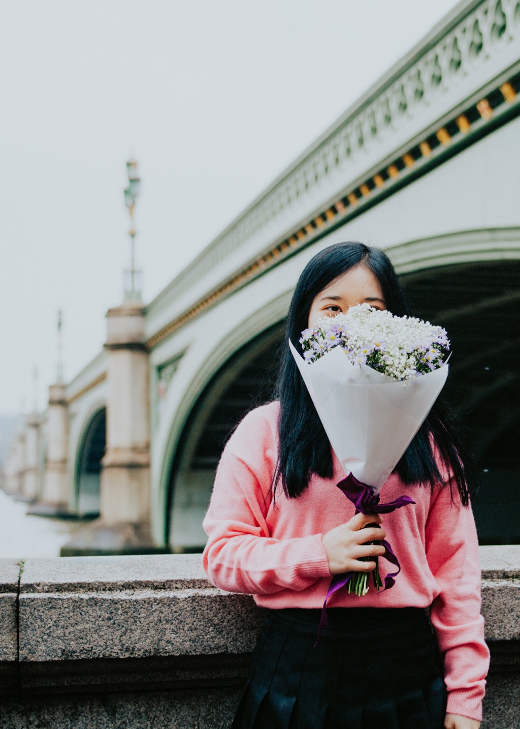 An au pair holds a bouquet of flowers while posing in front of a bridge with a view across the water to Big Ben in London. Conditions to au pair in the United Kingdom