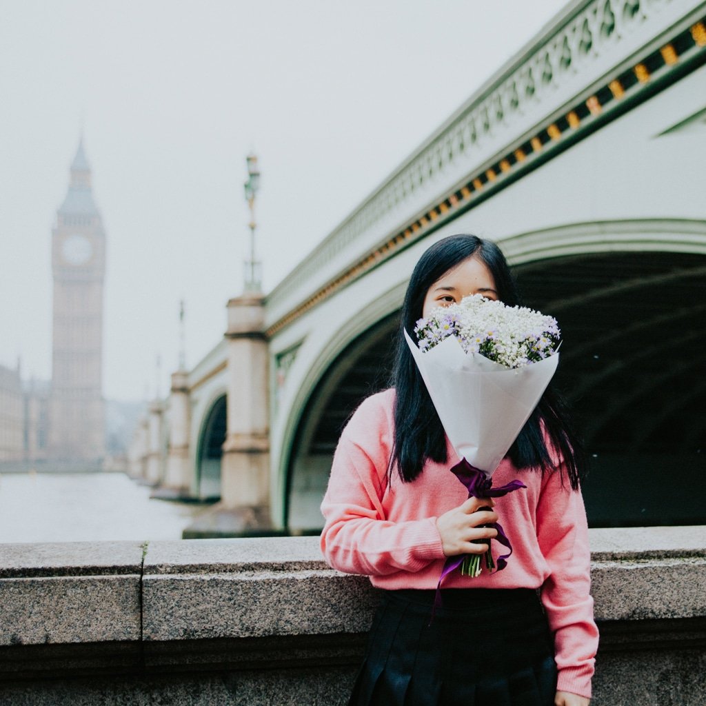 An au pair holds a bouquet of flowers while posing in front of a bridge with a view across the water to Big Ben in London.