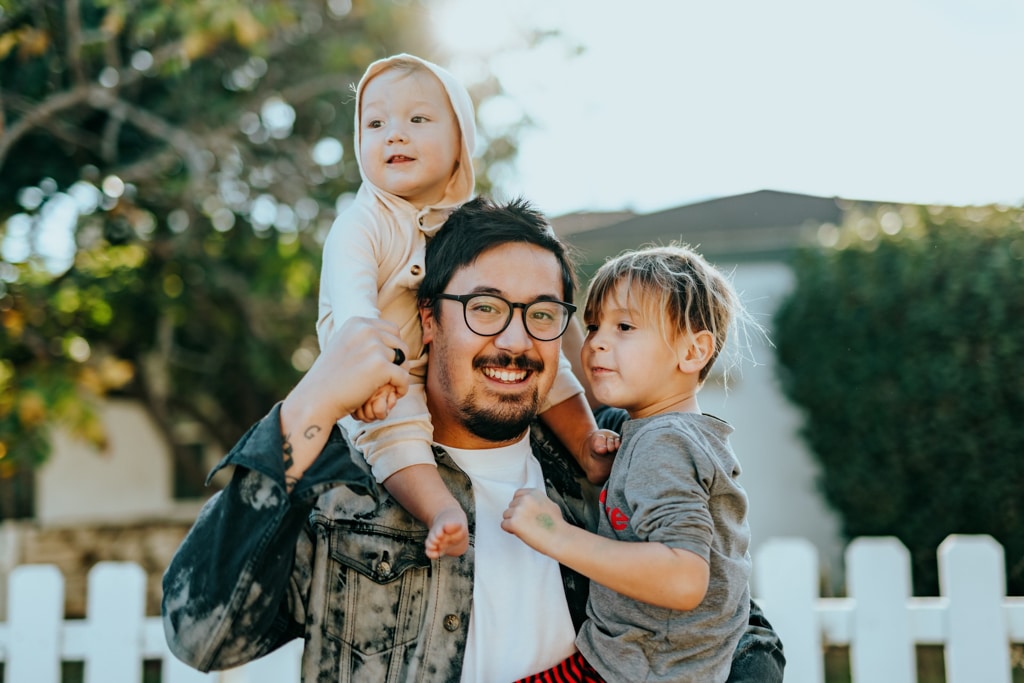 A father and his two children stand in front of their home and smile at the camera.