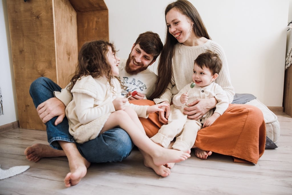 A family with two children sit together in the bedroom of their home.