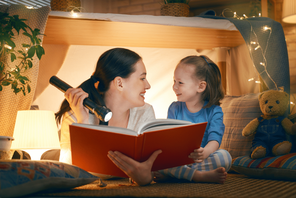 moeder ligt met haar kindje op de grond in een tent terwijl ze uit een boek leest met een zaklamp als onderdeel van het bedritueel
