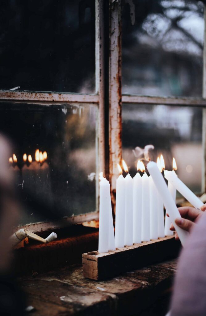 A hand holding a candle reaches out to light a menorah.