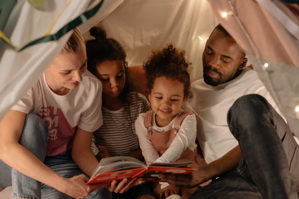 A family with two daughters reads a book in a fort they built in their home. Building a fort can be a fun game to play with your host children as it encourages their imagination and uses things you already have in the house!