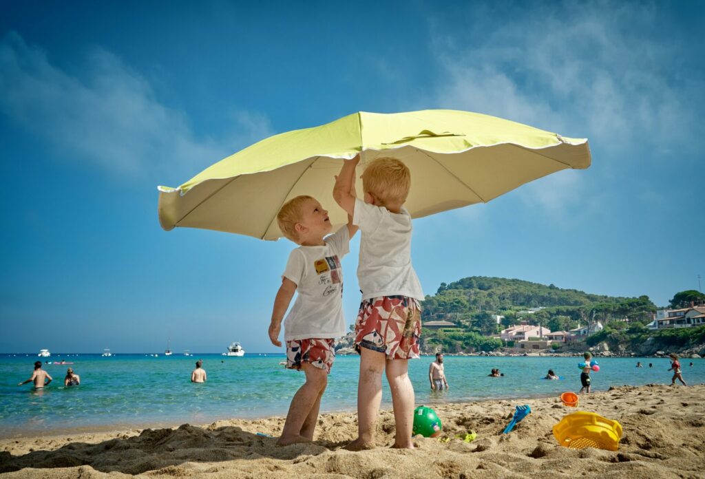 coronaproof op vakantie met kinderen. twee jongetjes met een wit shirt en rood, wit en blauwe zwembroek op het strand onder een gele parasol. 