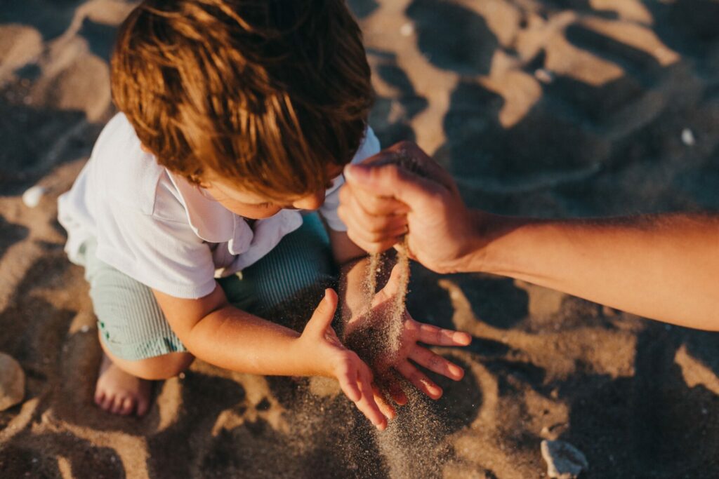 Kindje met wit shirt en groene broek op zijn hurken op het strand. Houdt zijn hand op terwijl iemand anders er zijn in gooit. 