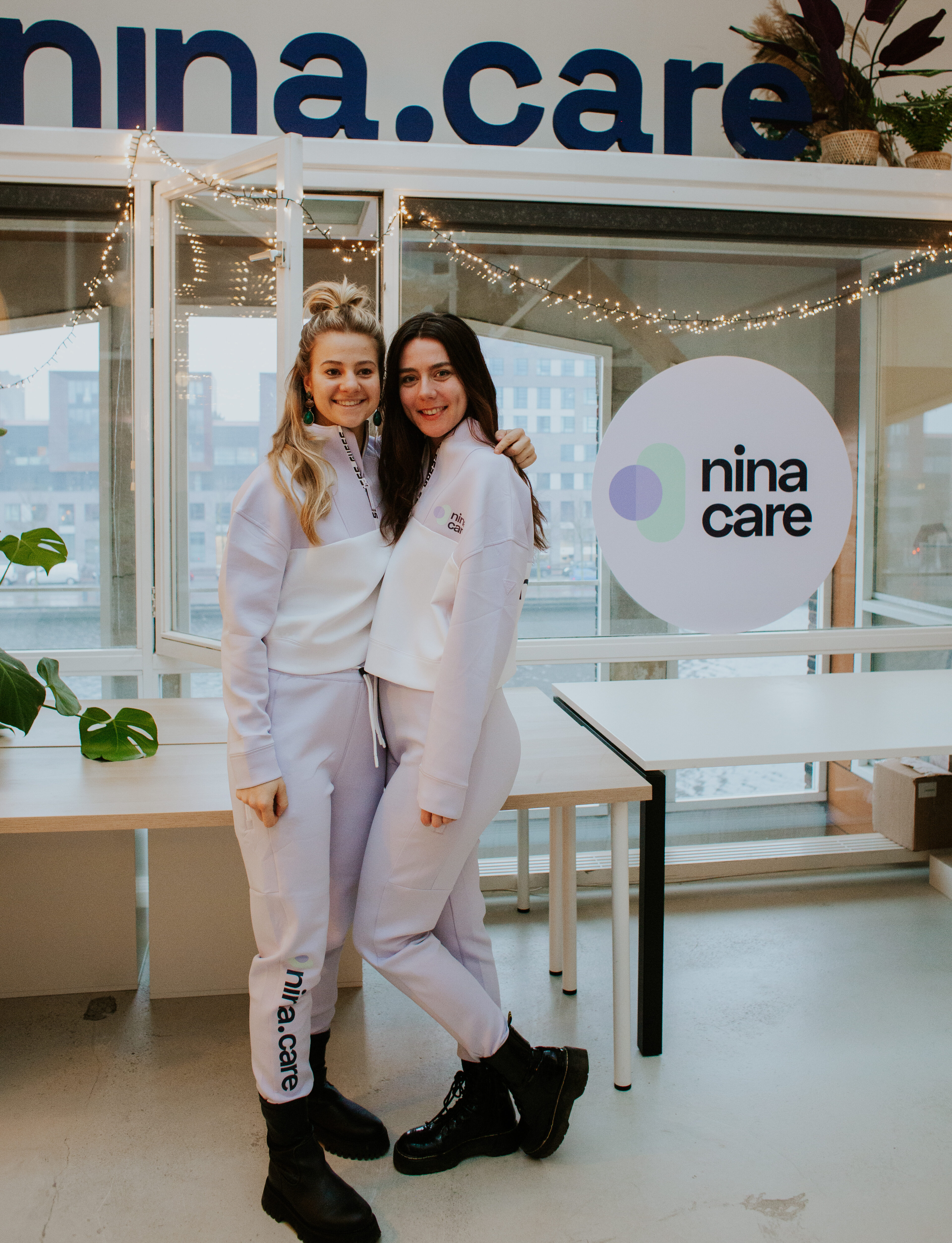 twee meiden die het nina.care uniform showen door te poseren onder het nina.care logo op kantoor